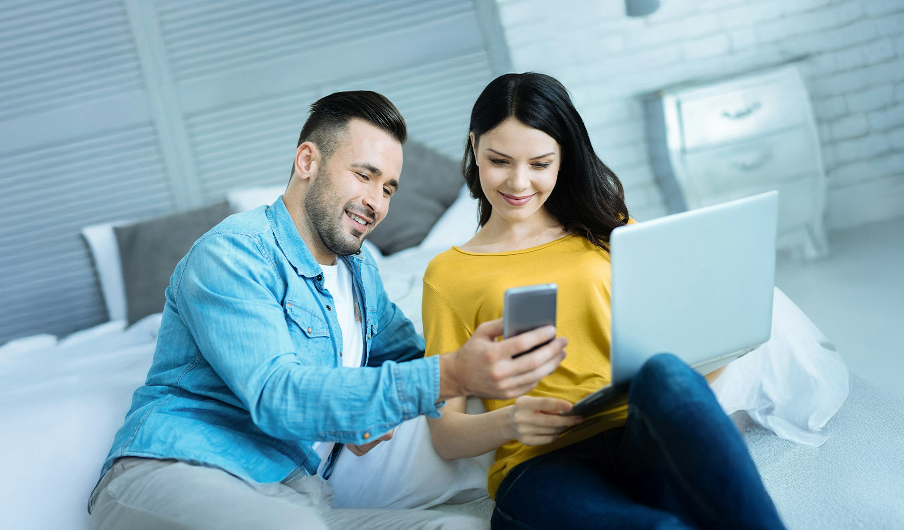 Ein glückliches, junges Pärchen, sitzt in einem hellen Raum, am Laptop und Handy. Golden Gates biete auch für Sie den passenden Sparplan um Ihr Geld in physische Edelmetalle anzulegen.